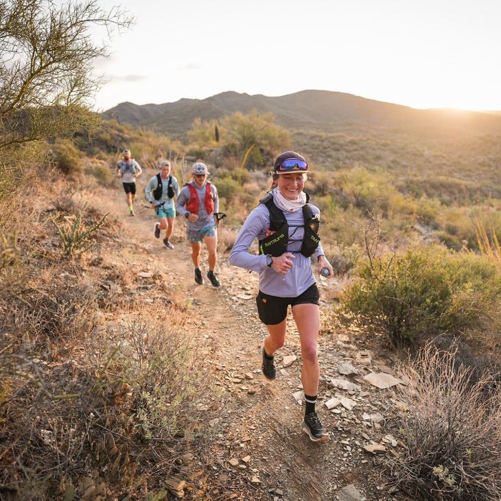 Four runners pass by a cactus on a desert trail at sunrise.