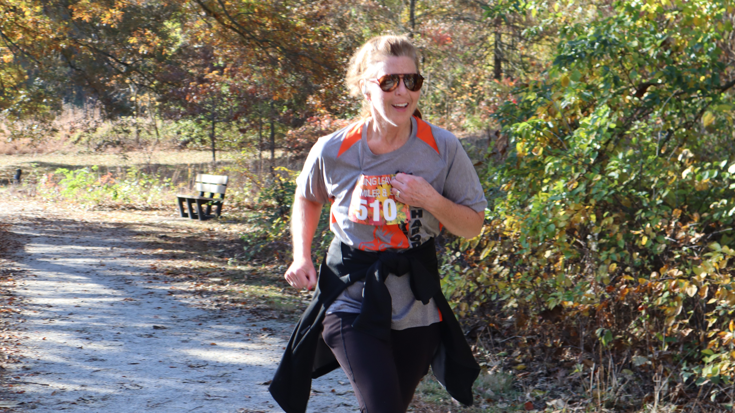 Woman in running clothes, sunglasses and racing bib runs on trail with trees in fall colors in the background.
