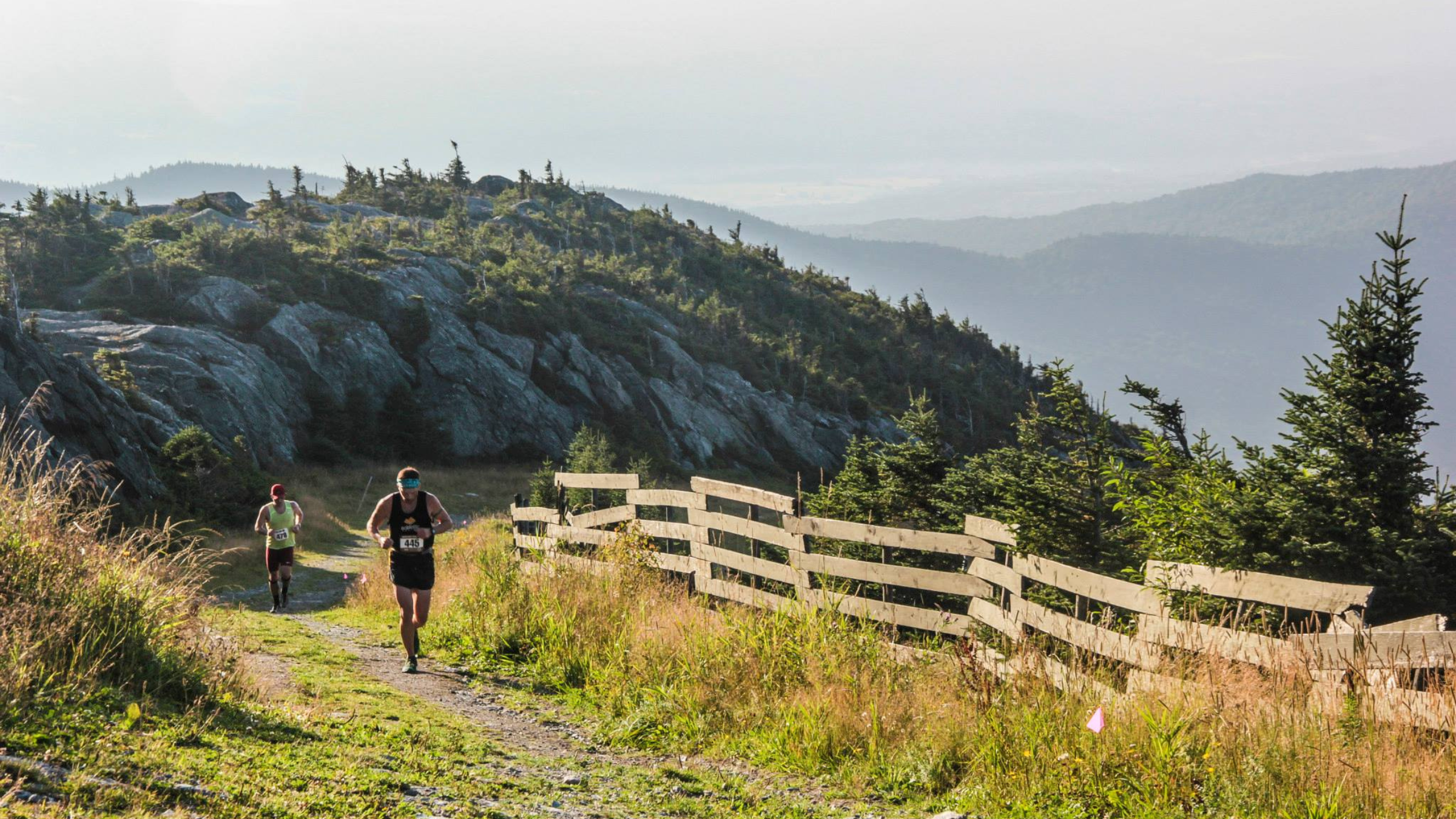 Two men with race bibs on shirts run on trail toward camera alongside wooden fence with evergreen trees and multiple mountains in the background.