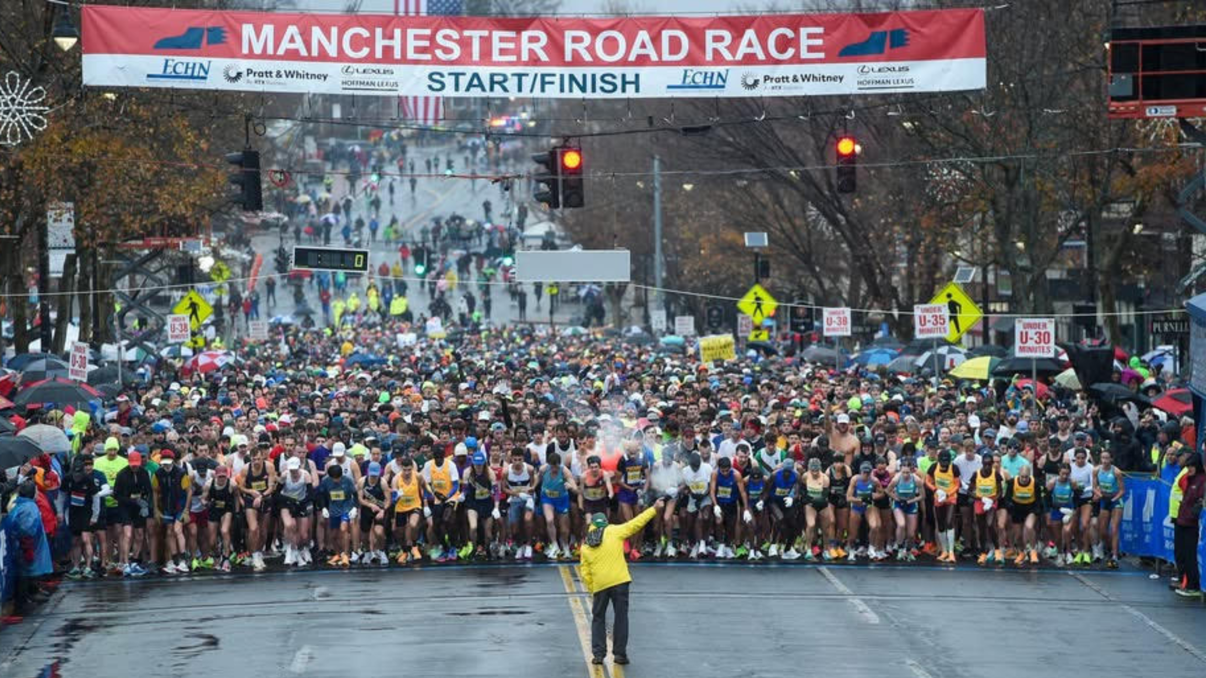 thousands of runners line up to start off-distance race on street with race official facing them to shoot off official gun, under a banner that reads "Manchester Road Race Start/Finish"