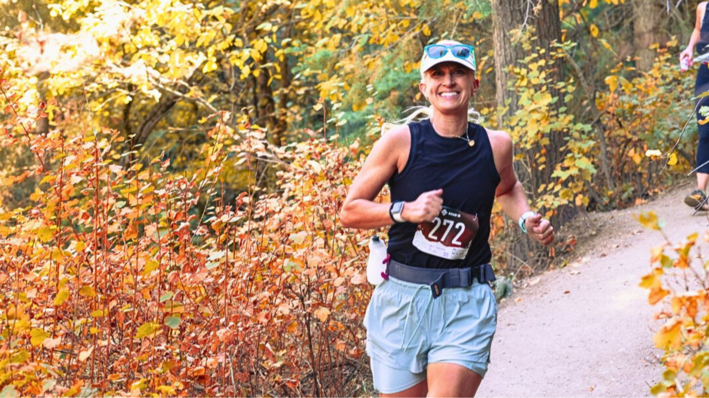 Woman with race bib on shirt runs on trail along bushes and trees in fall colors.