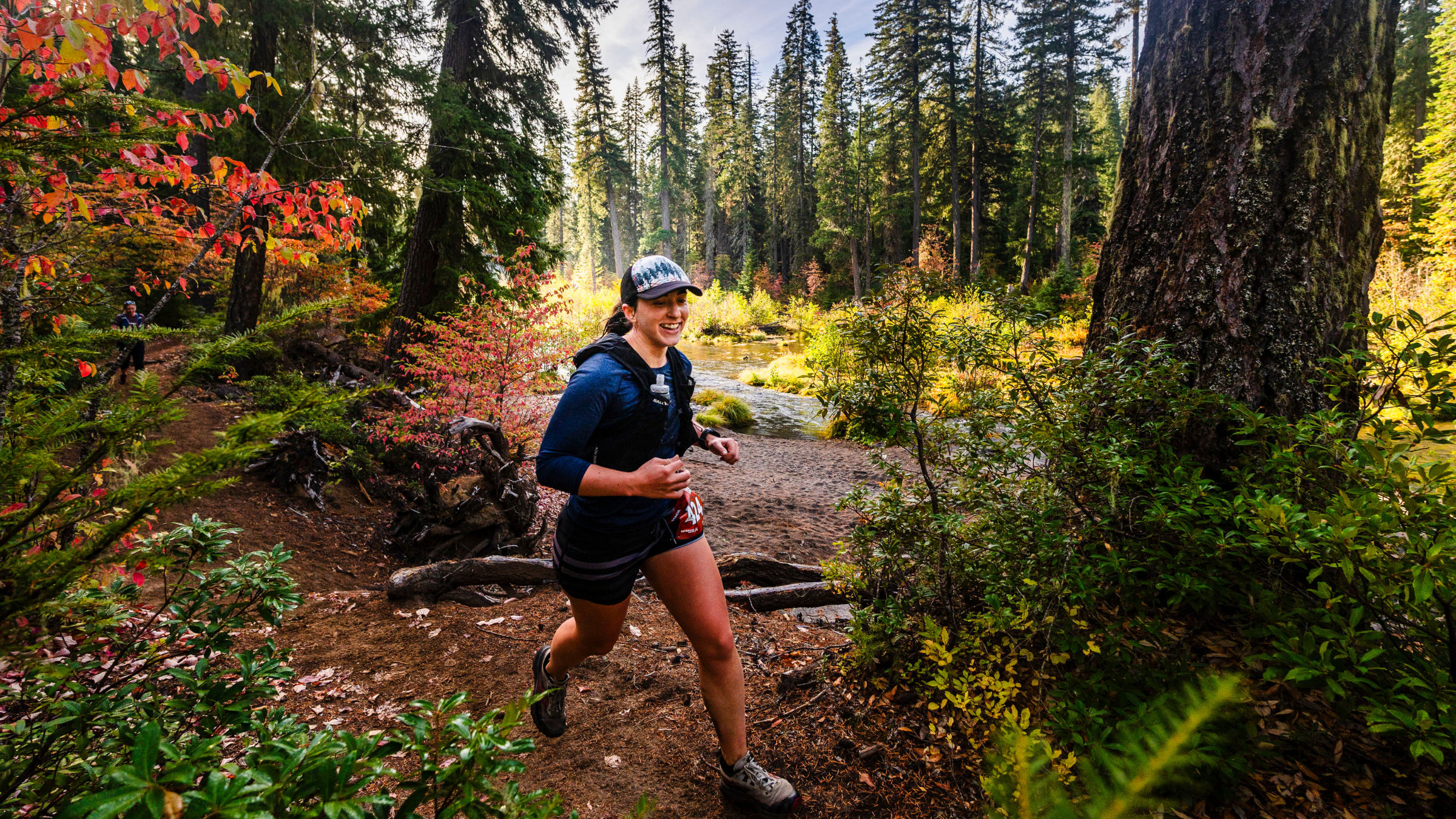 Woman runs on trails with hydration pack and hat on with evergreen trees and fall foliage surrounding her.
