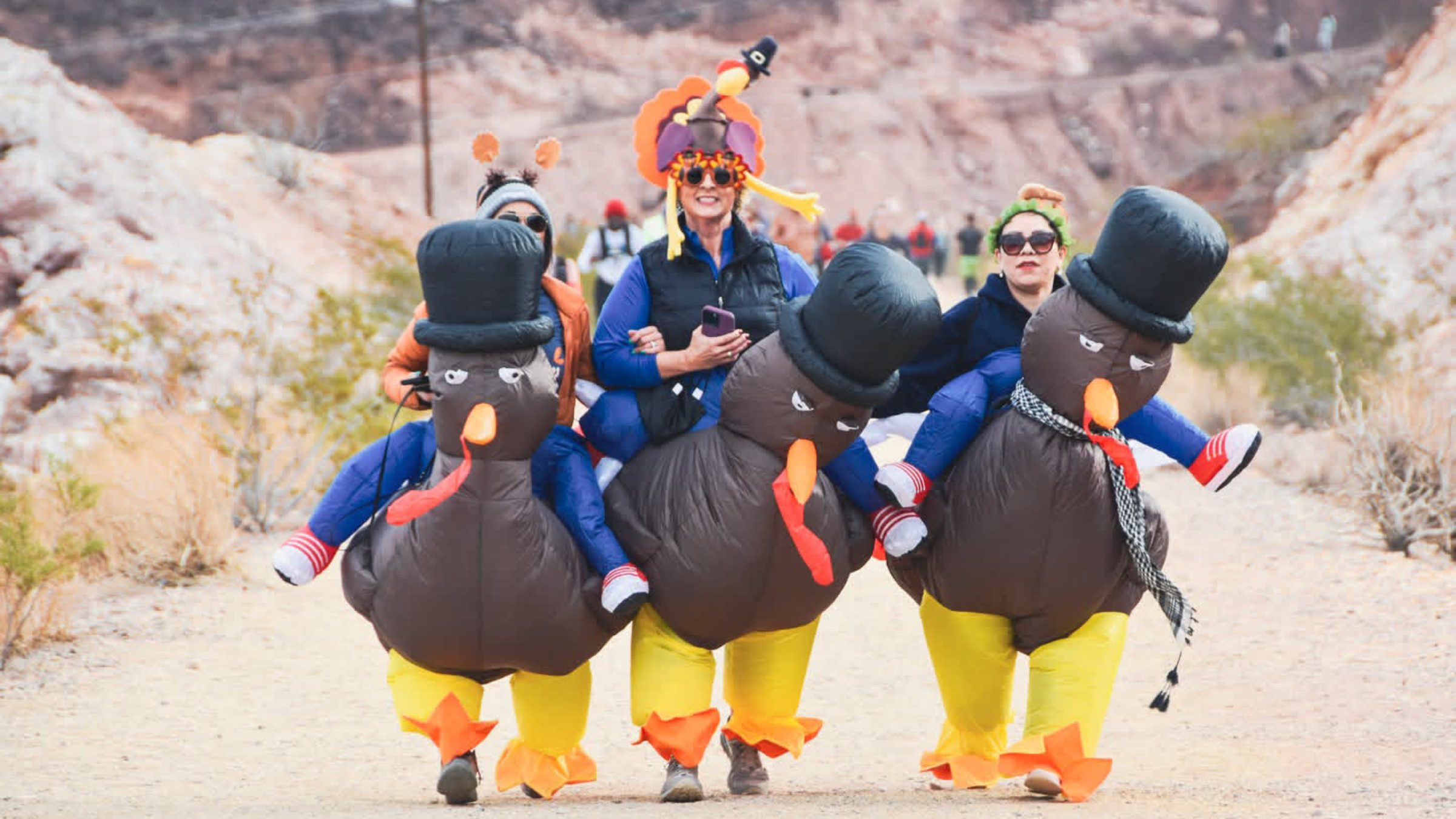 three runners in inflatable turkey costumes run on a road within in a desert landscape