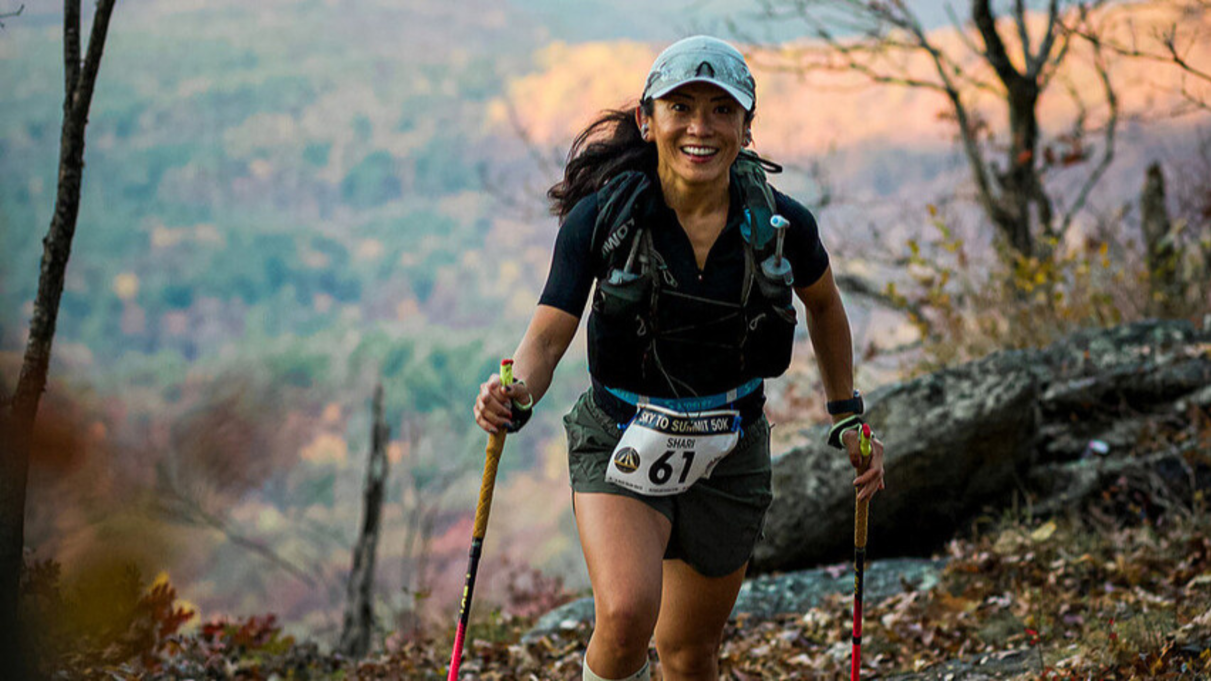 Middle-aged woman in running gear with hat and backpack and race bib and trekking poles walks toward camera on trail with trees and valley views behind her.