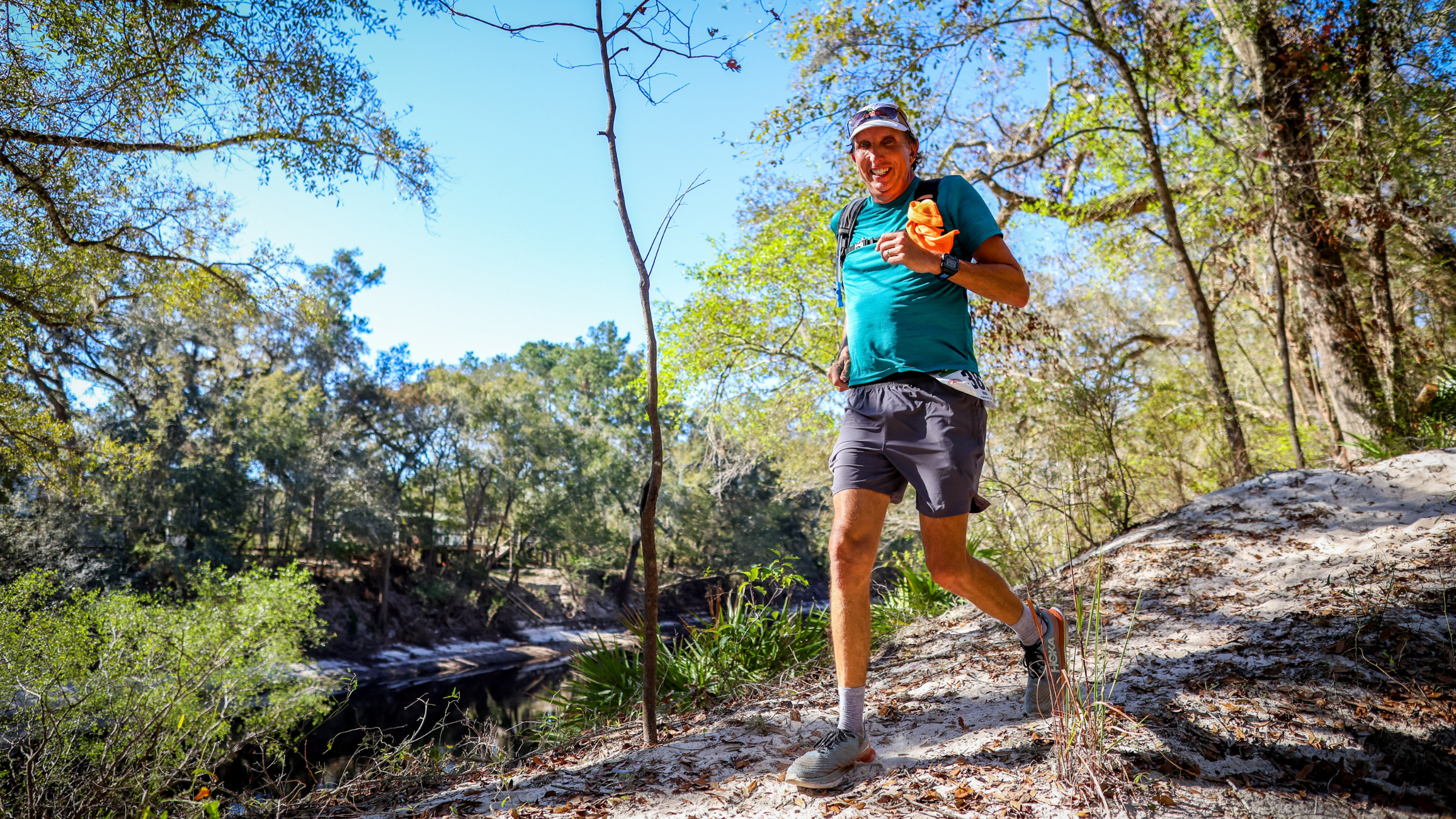 Middle-aged man with hydration pack and running clothes on runs downhill on trail with trees and river in the background.
