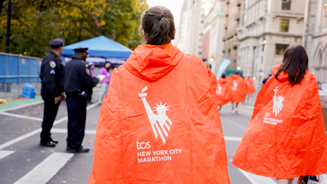 women wearing orange NYC Marathon ponchos walk away from the camera on road with police officers in the background